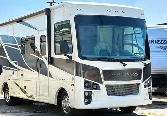 White Class A motorhome parked in a lot, with a large front windshield, chrome grille, side mirrors, and deployed entry steps.
