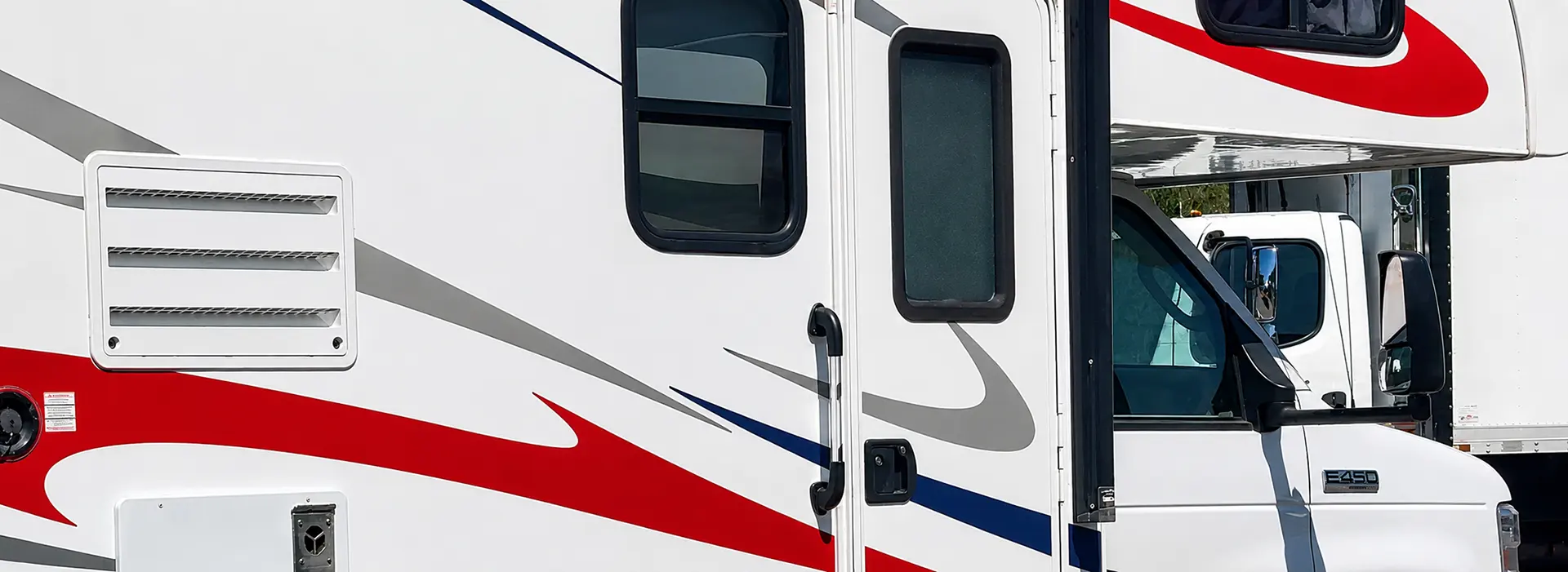 White motorhome with red and gray swoosh graphics, open entry door, and side window; another truck is visible in the background.